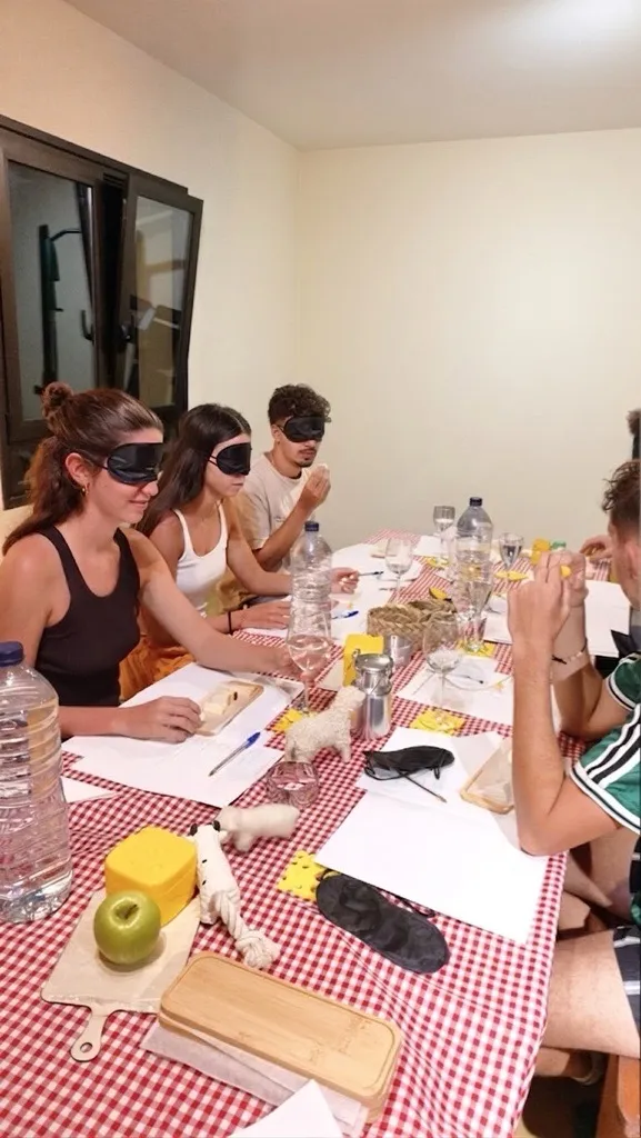 Un grupo de asistentes con antifaz cata quesos a ciegas alrededor de una mesa con mantel de cuadros rojos, con fichas de cata y copas de agua.
