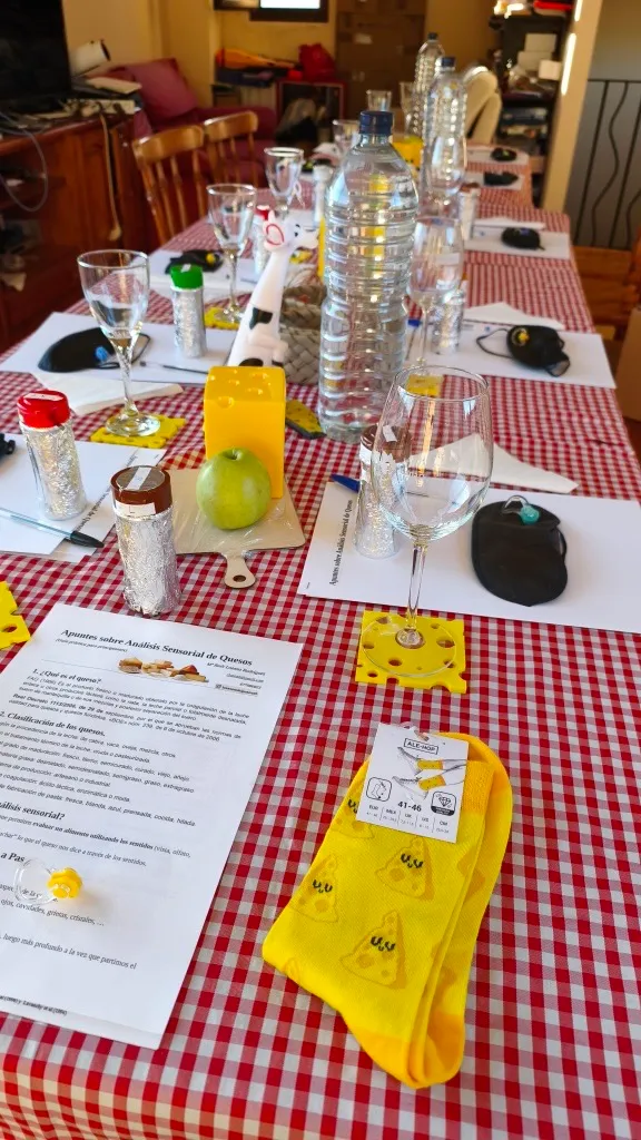 Detalle de una mesa de cata: apuntes sobre análisis sensorial de quesos, tarros de aromas envueltos en papel de aluminio, antifaces, manzana verde y copas.